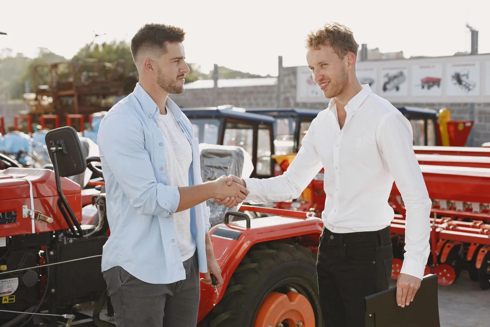 Two people shaking hands beside farm equipment, sealing a business equipment purchase