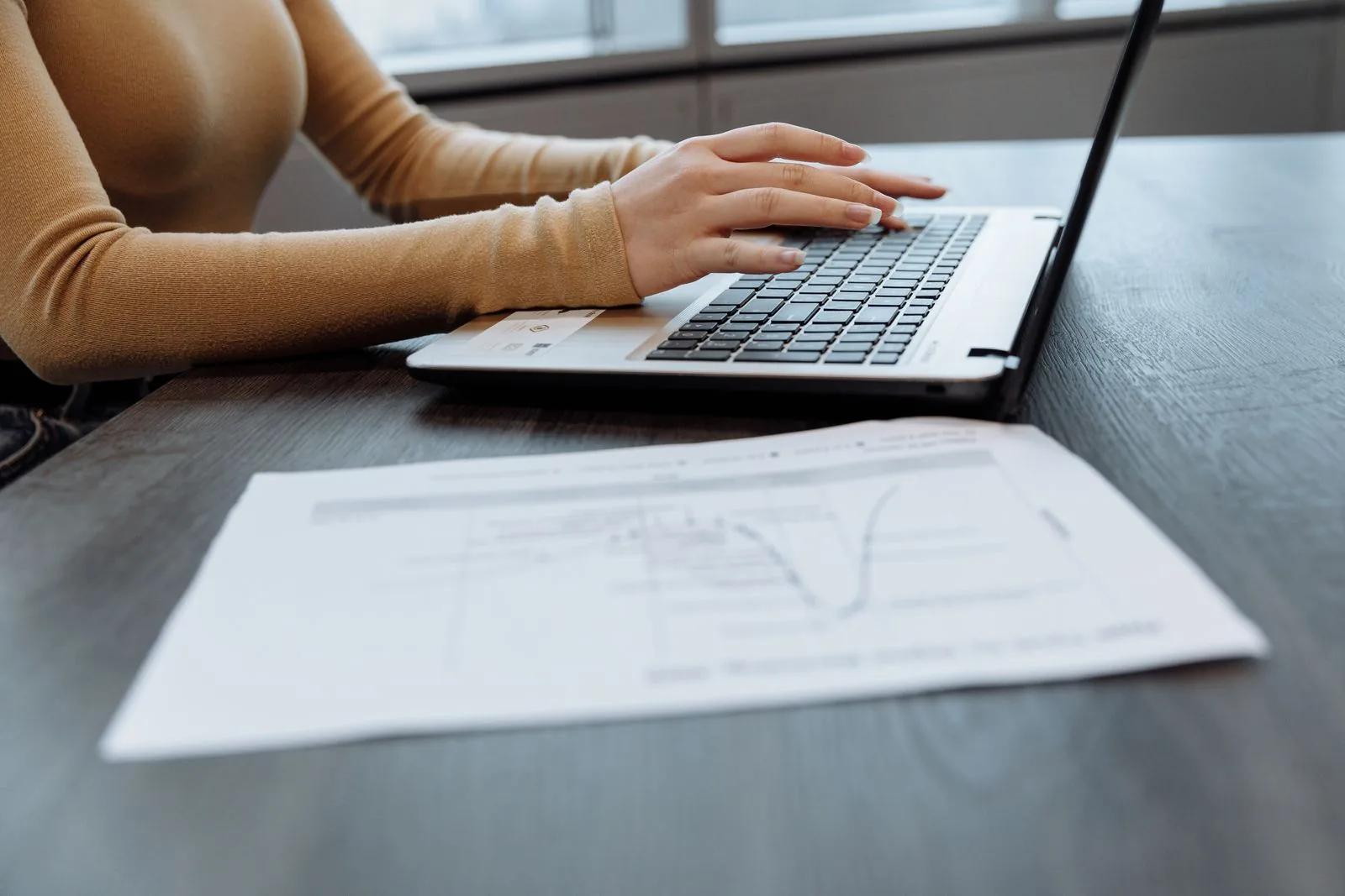 Close-up of a contractor's hands typing on a laptop beside paperwork at a desk