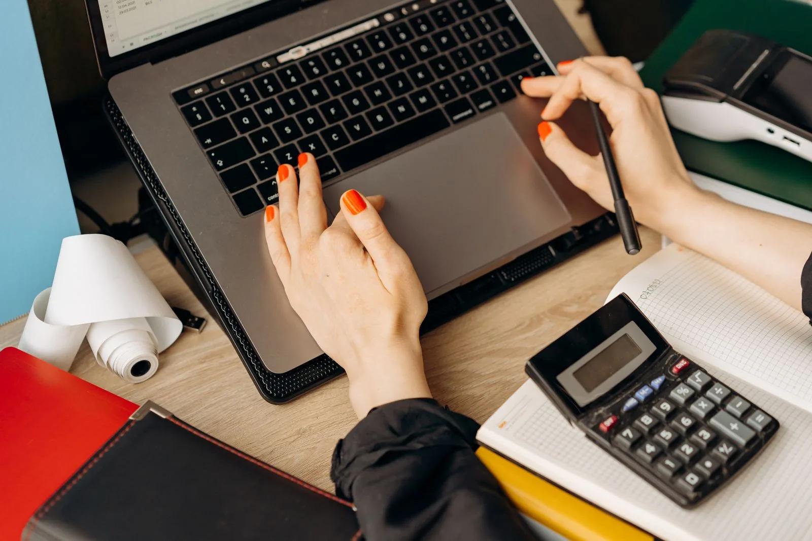 A person using a laptop and calculator on a wooden desk for bookkeeping