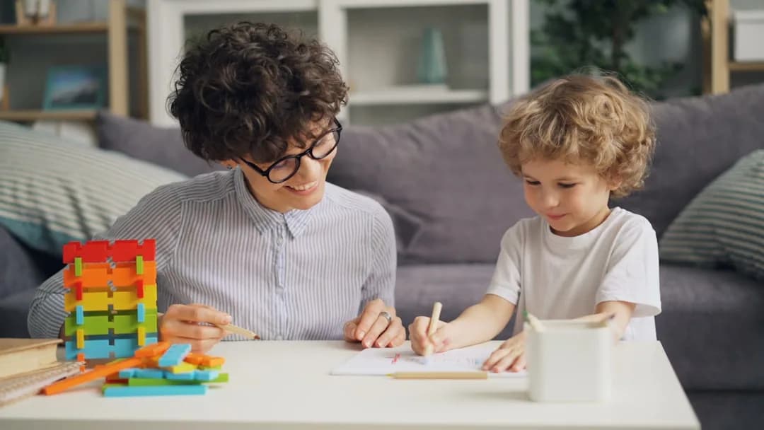 A childminder at a kitchen table reviewing fees and receipts on a laptop while children play nearby