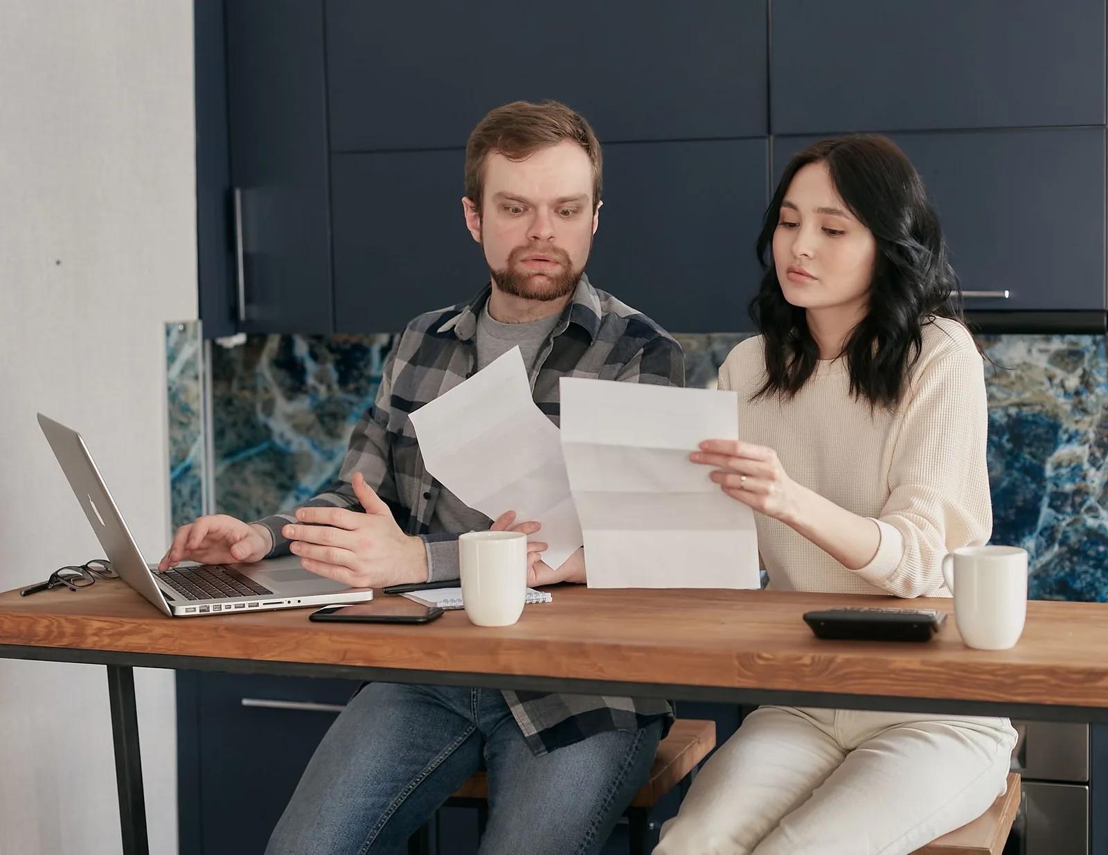 A couple sitting at a kitchen table going through household bills and paperwork with a laptop and calculator
