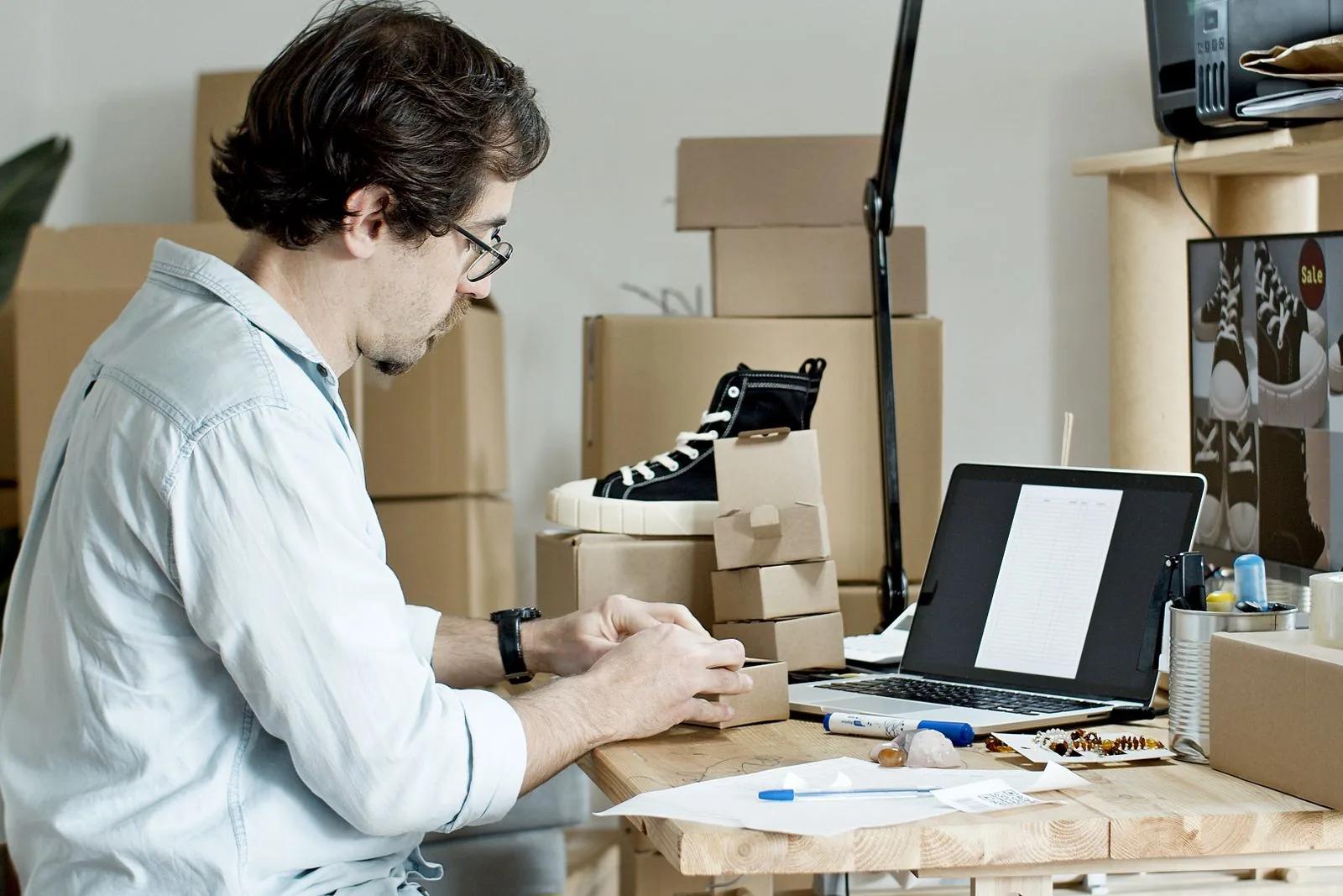 Ecommerce seller packing product orders at a desk with laptop and boxes