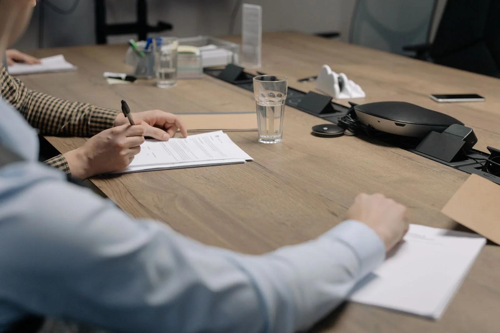 Hands signing official company documents at a wooden meeting table