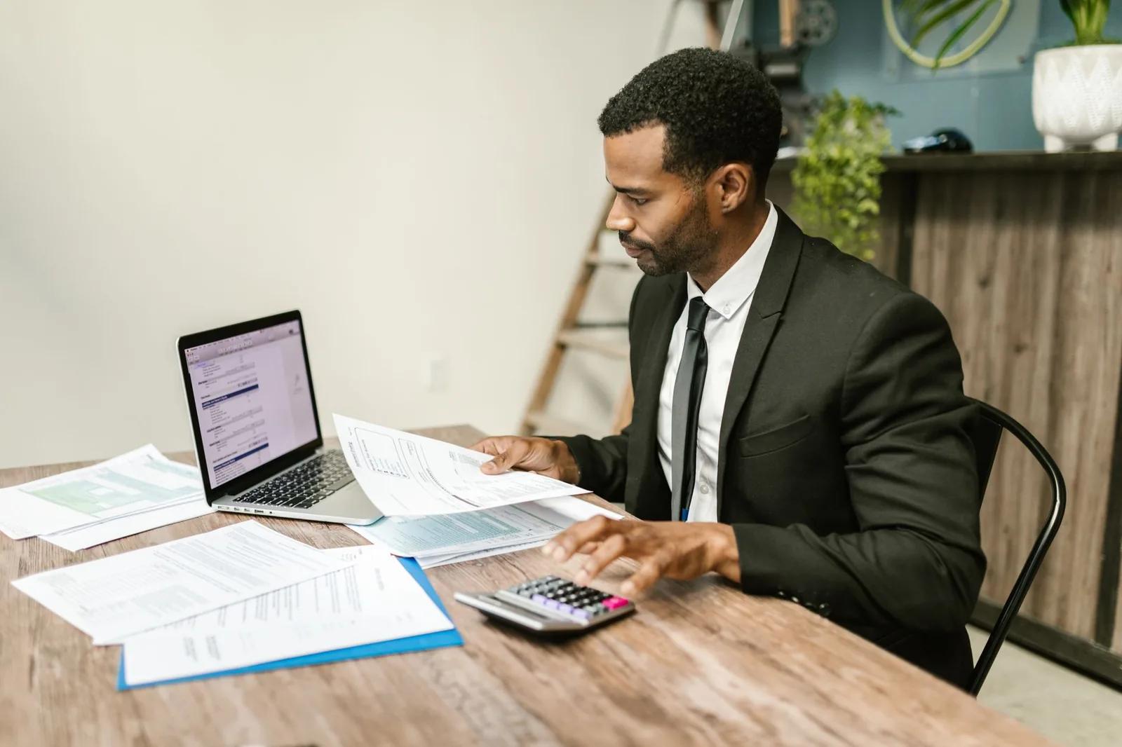 A UK company director reviewing business finances and tax payments on a laptop at a desk, with documents and a calculator
