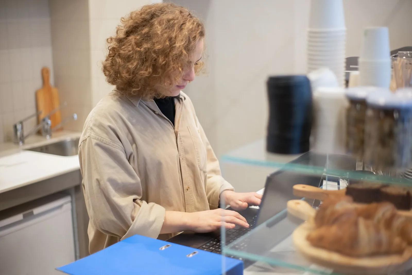 A UK cafe owner checking staff rota and payroll figures on a laptop at the counter of a bright small business