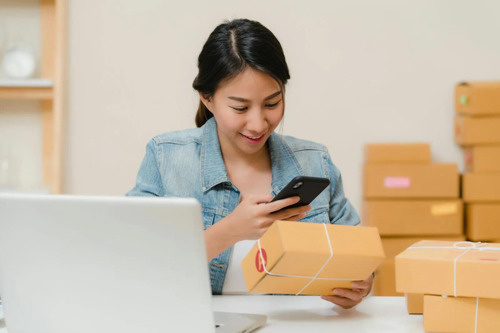 A small online seller checking an order on her phone at a laptop desk, surrounded by parcels ready to post