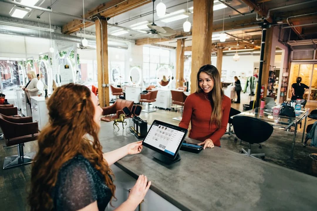 A salon owner reviewing staffing costs at the front desk while a stylist works at a chair in the background
