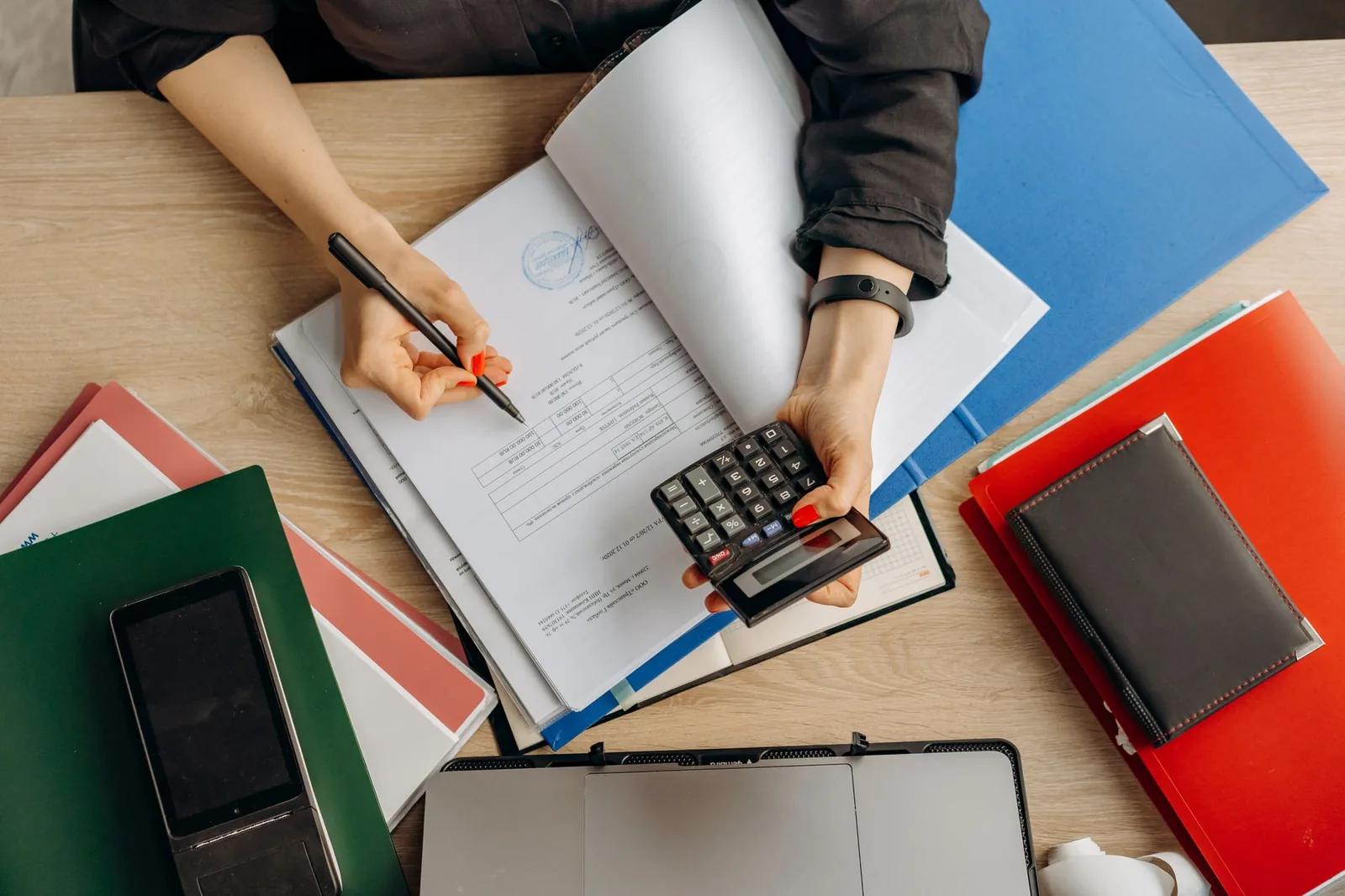 An accountant using a calculator over an open accounts ledger, with folders and a laptop on the desk
