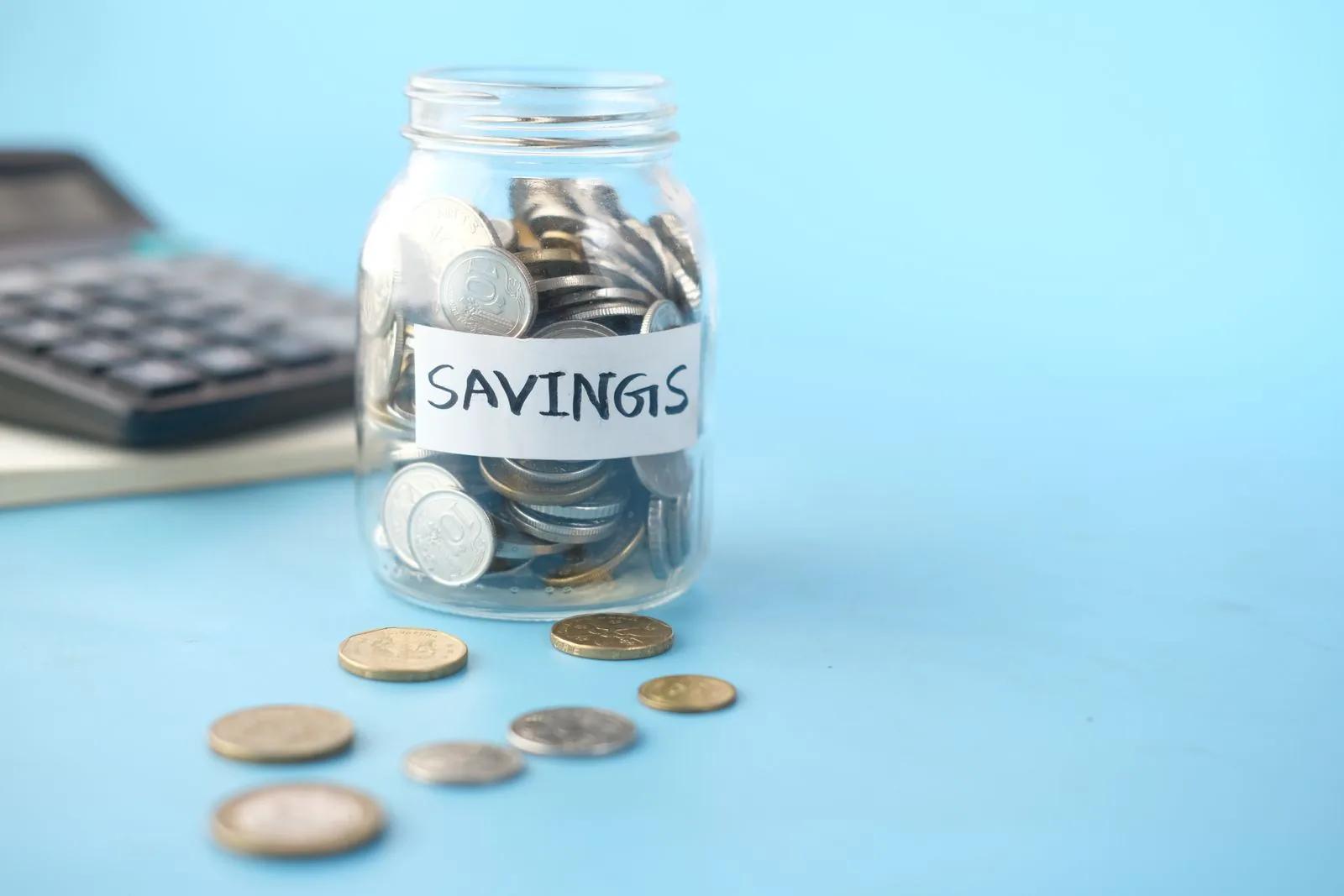 Glass jar of coins labelled savings beside a calculator on a blue background