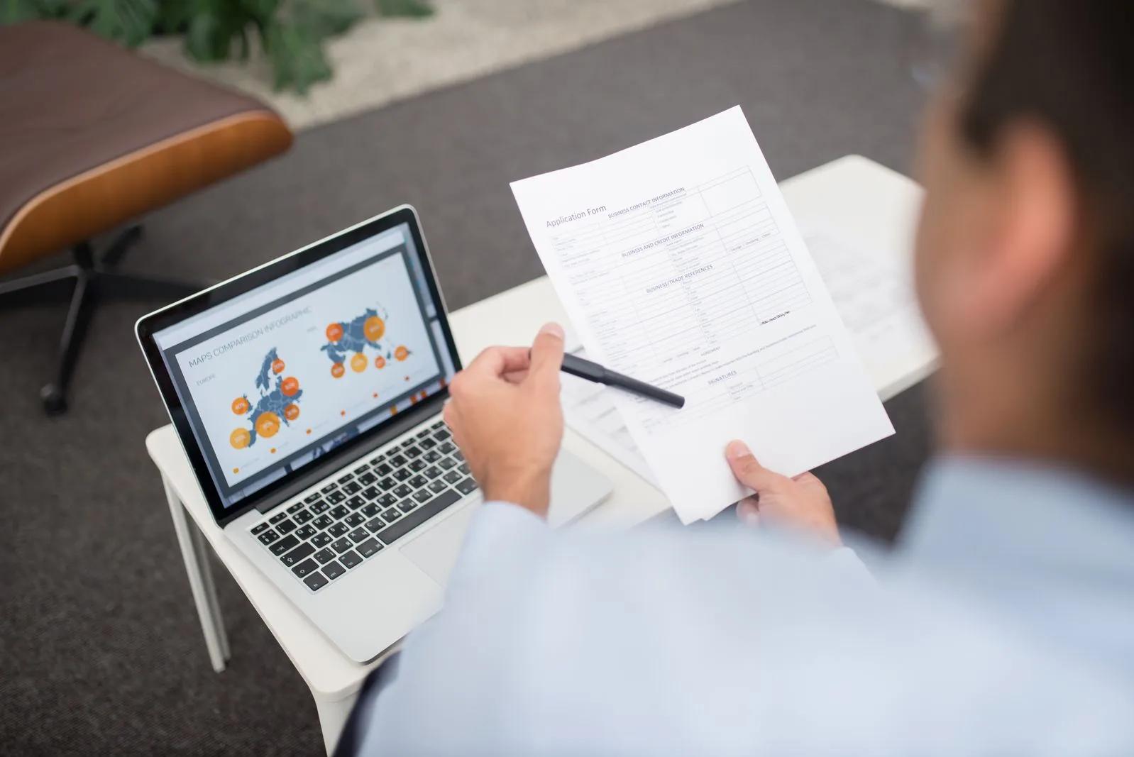 A business owner reviewing documents and data on a laptop while completing VAT registration paperwork