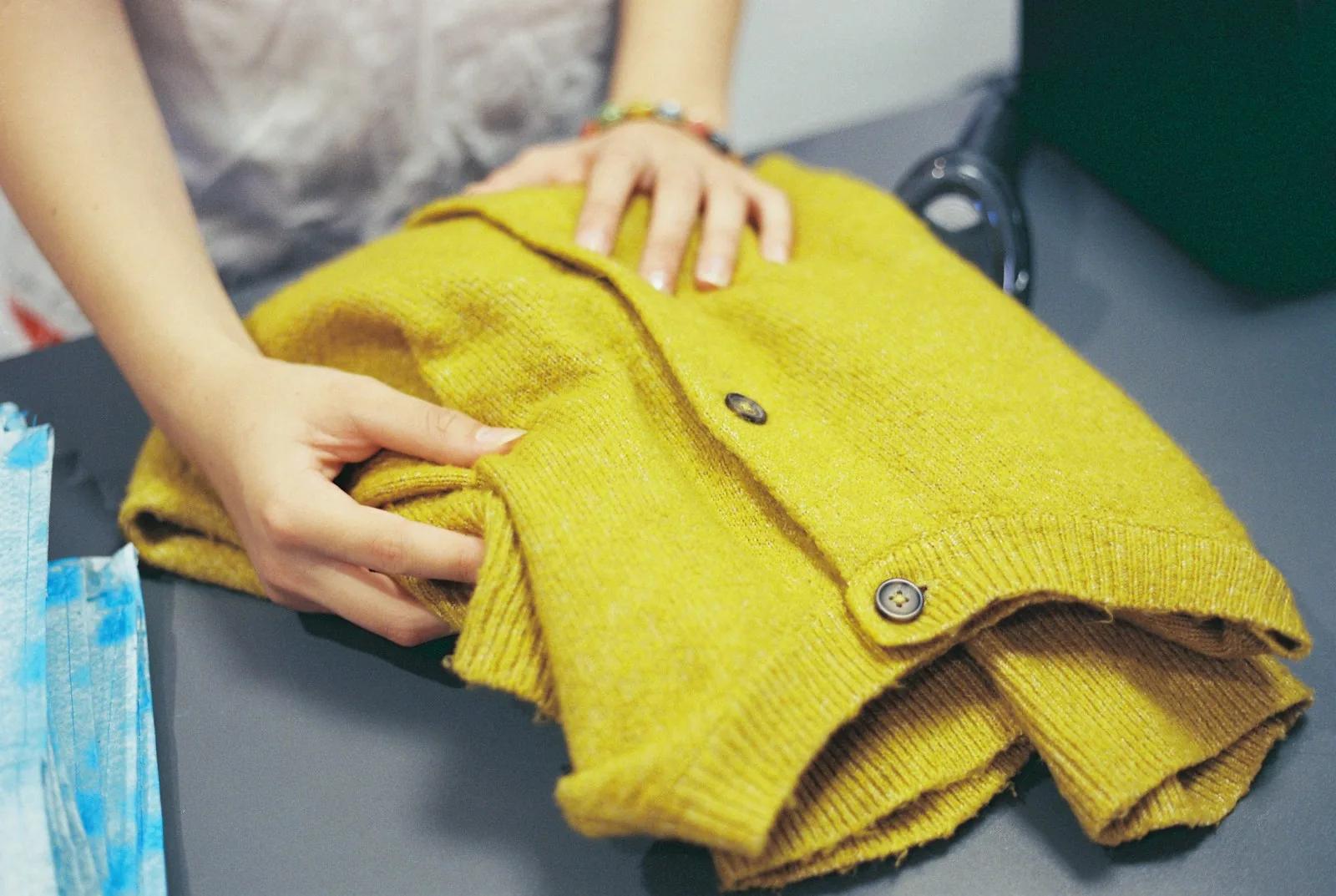 Hands folding a mustard-yellow knitted cardigan on a grey counter before it is sold on
