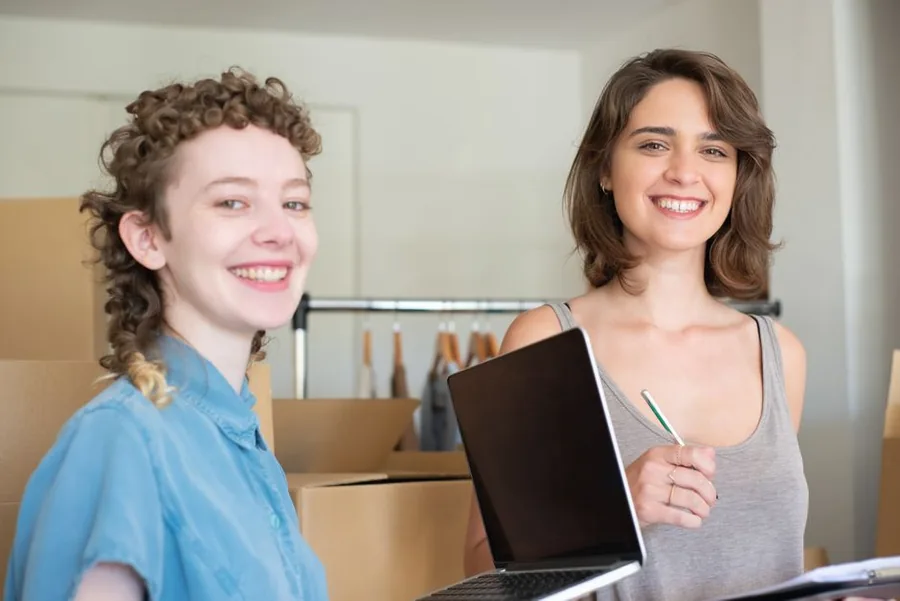 Two online store owners reviewing sales figures on a laptop beside stock boxes ready to ship to customers Two online store owners reviewing sales figures on a laptop beside stock boxes ready to ship to customers
