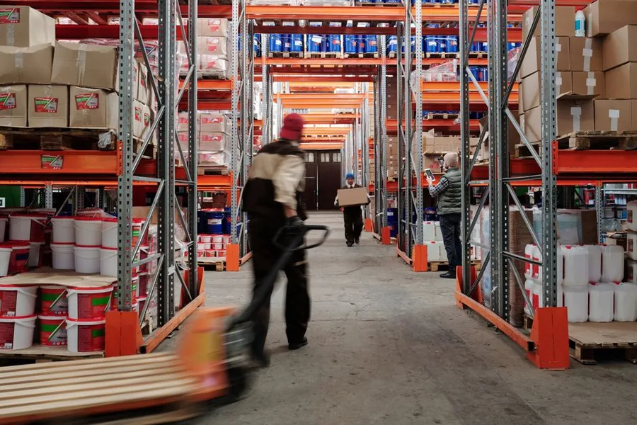 Aisle of tall warehouse racking packed with stock and pallets, the kind of long-held inventory that triggers Amazon FBA storage fees Aisle of tall warehouse racking packed with stock and pallets, the kind of long-held inventory that triggers Amazon FBA storage fees