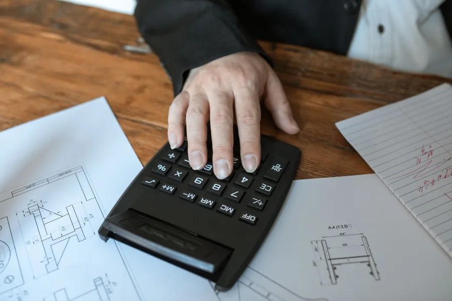 A construction contractor working out figures on a calculator over technical drawings, calculating tax due on retention held back by clients A construction contractor working out figures on a calculator over technical drawings, calculating tax due on retention held back by clients
