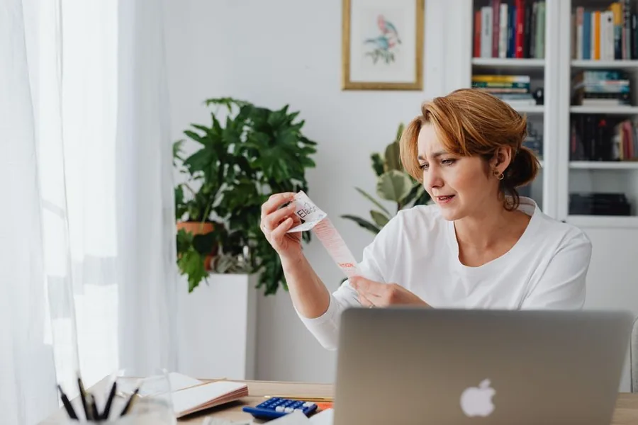 A sole trader at her home desk checks a receipt against her laptop while totting up allowable business expenses A sole trader at her home desk checks a receipt against her laptop while totting up allowable business expenses
