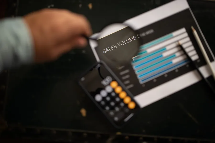 A magnifying glass held over a printed sales volume chart beside a calculator, reviewing ecommerce advertising performance A magnifying glass held over a printed sales volume chart beside a calculator, reviewing ecommerce advertising performance