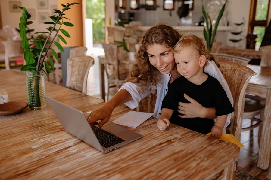 A childminder catching up on her accounting software at the kitchen table while keeping an eye on a young child A childminder catching up on her accounting software at the kitchen table while keeping an eye on a young child