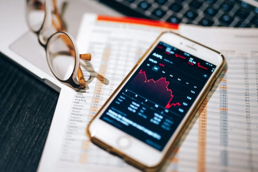 A share portfolio tracked on a smartphone stock app beside reading glasses and a printed market performance table