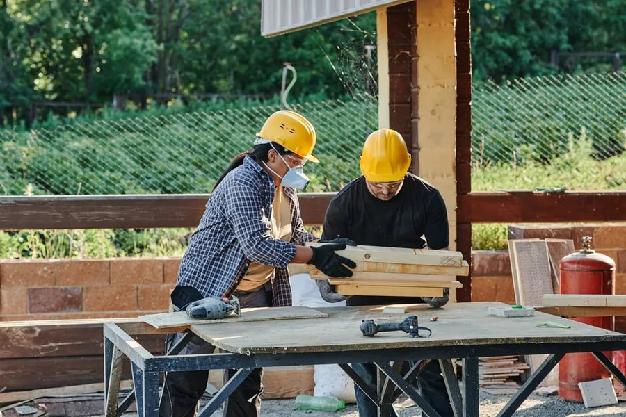 Two builders in hard hats handling timber on site, the kind of materials and labour split that matters under CIS Two builders in hard hats handling timber on site, the kind of materials and labour split that matters under CIS
