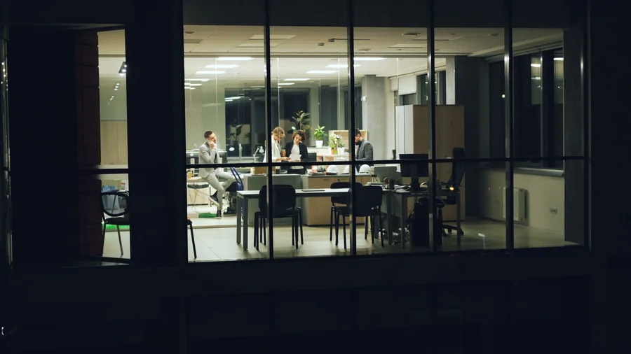 A city-centre office lit up at night with staff working late inside, seen from outside A city-centre office lit up at night with staff working late inside, seen from outside
