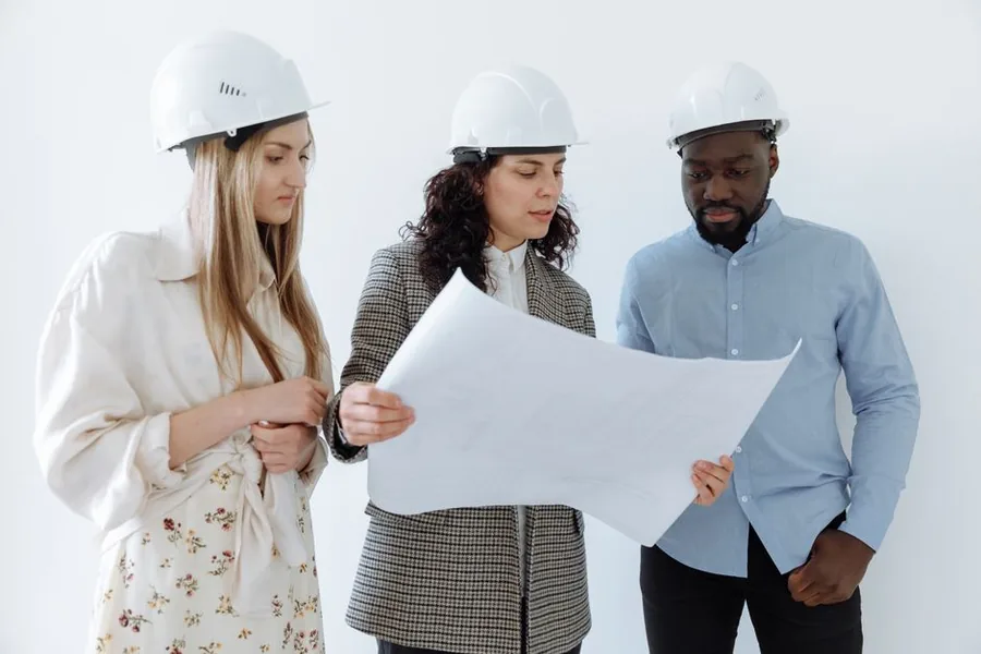 Consulting engineers in hard hats reviewing technical drawings together while planning a project Consulting engineers in hard hats reviewing technical drawings together while planning a project