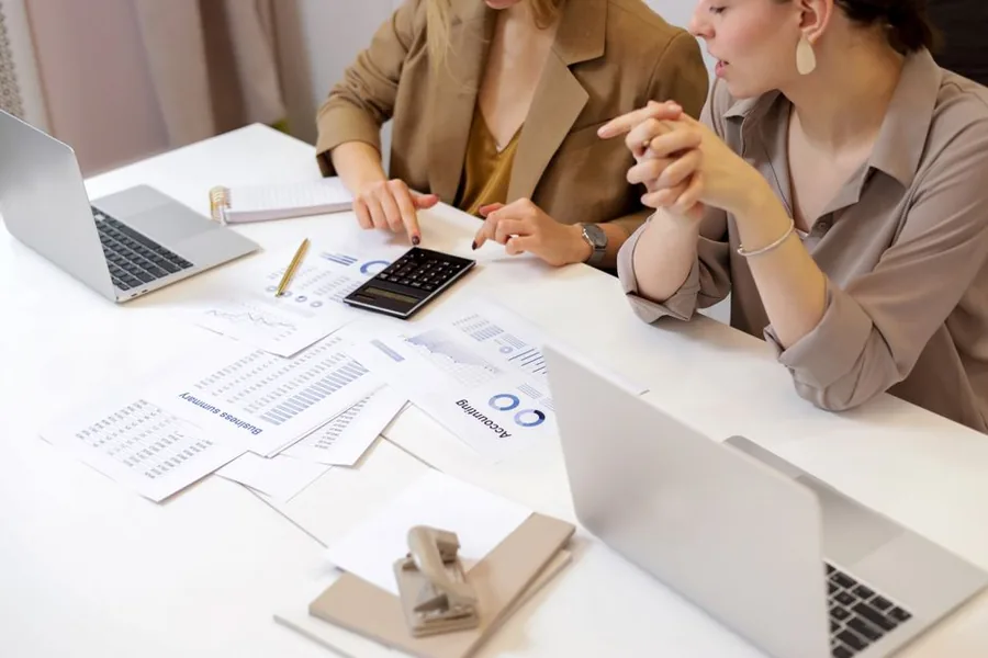 Two creative agency colleagues reviewing financial summaries and cashflow figures together over laptops and a calculator Two creative agency colleagues reviewing financial summaries and cashflow figures together over laptops and a calculator