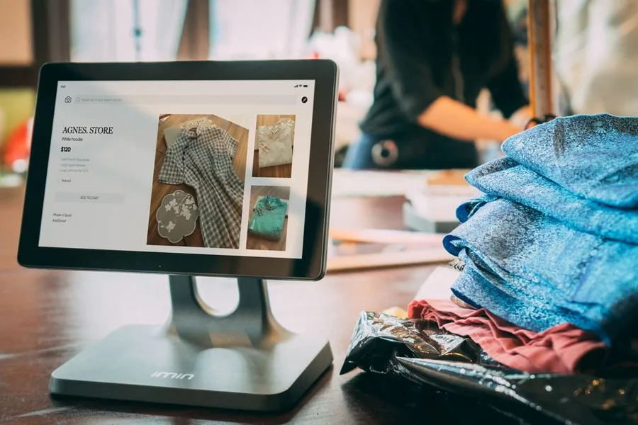 A UK online clothing seller checking a product listing on a point-of-sale screen beside folded stock ready to pack and dispatch A UK online clothing seller checking a product listing on a point-of-sale screen beside folded stock ready to pack and dispatch