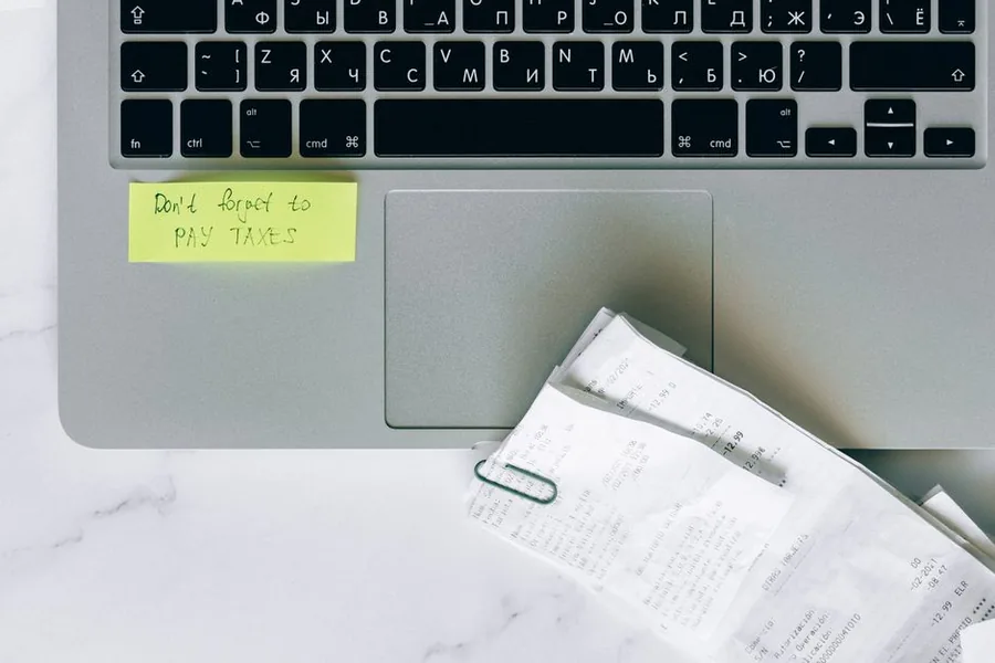A pay taxes reminder note stuck to a laptop beside a bundle of paper receipts on a desk A pay taxes reminder note stuck to a laptop beside a bundle of paper receipts on a desk