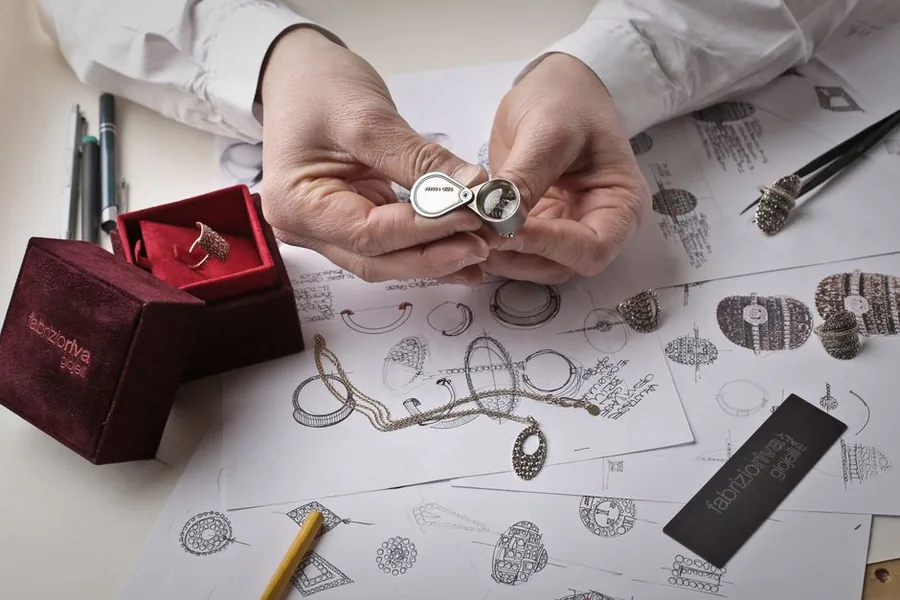A jeweller examining a finished piece through a loupe beside hand-drawn ring designs at the bench A jeweller examining a finished piece through a loupe beside hand-drawn ring designs at the bench
