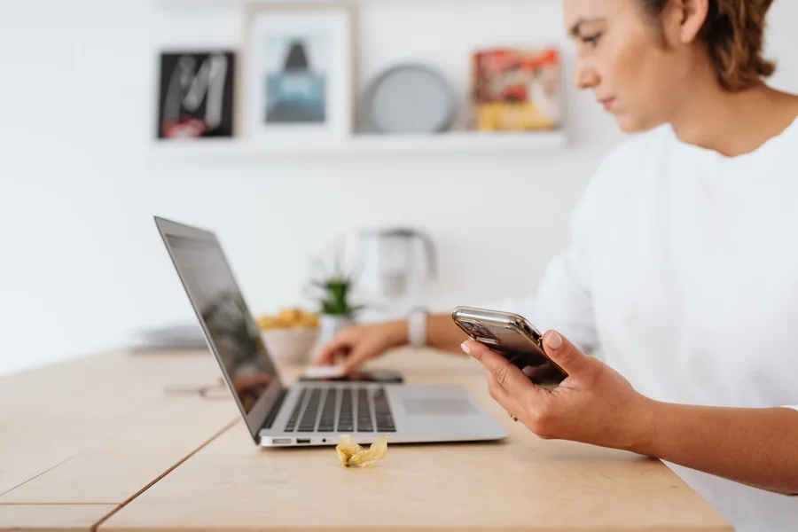 Small business owner checking her smartphone while typing on a laptop at a bright home office desk, an everyday secure sign-in moment. Small business owner checking her smartphone while typing on a laptop at a bright home office desk, an everyday secure sign-in moment.