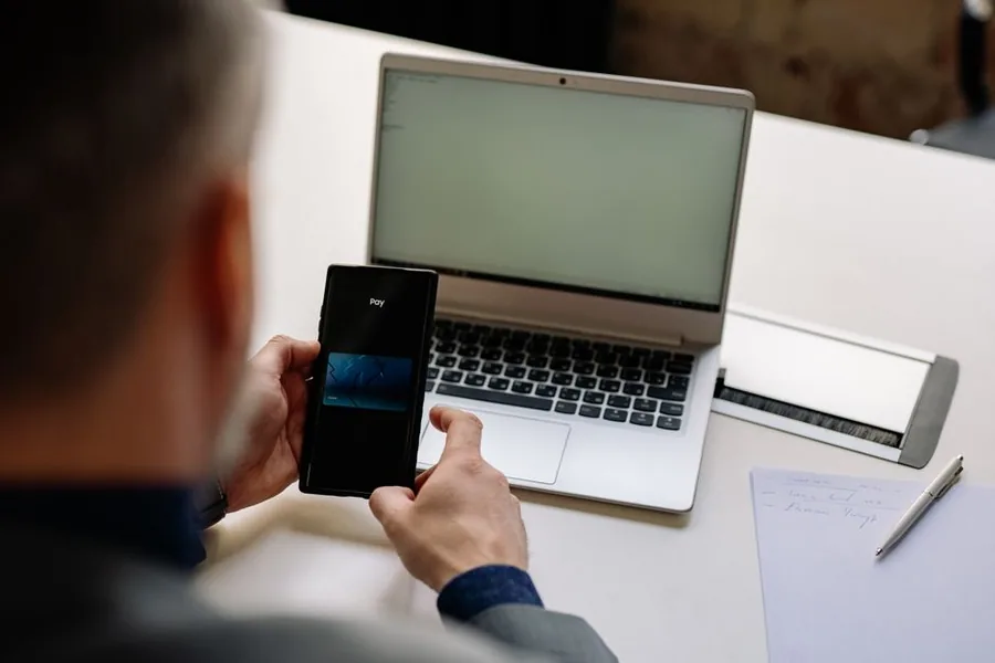 A company director paying a tax bill online, using a phone banking app alongside a laptop at their desk A company director paying a tax bill online, using a phone banking app alongside a laptop at their desk