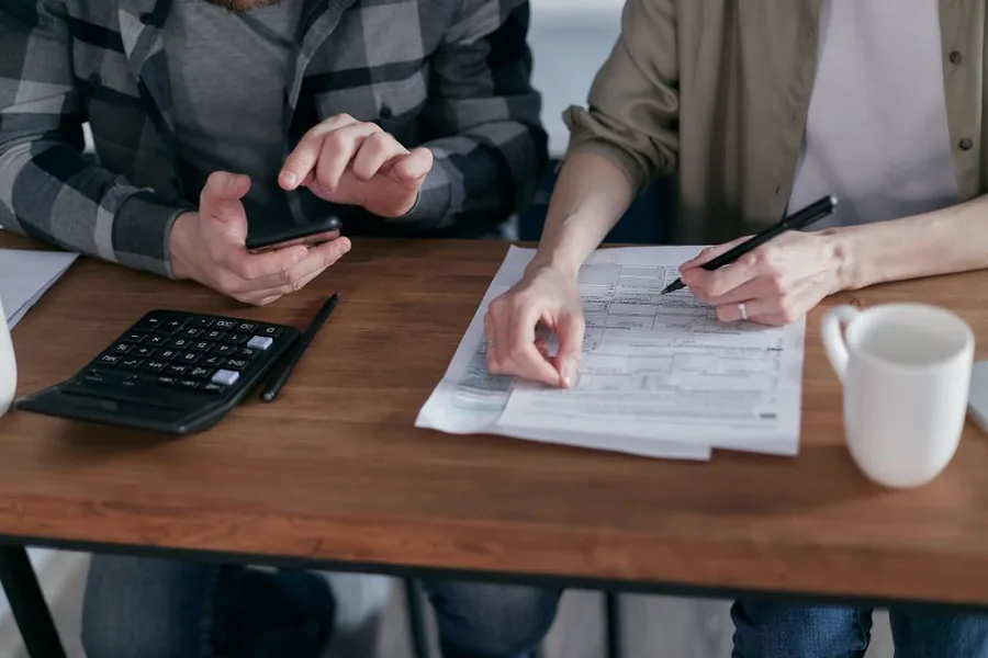 A couple at a kitchen table checking paperwork and a calculator while updating their personal details after a name change A couple at a kitchen table checking paperwork and a calculator while updating their personal details after a name change