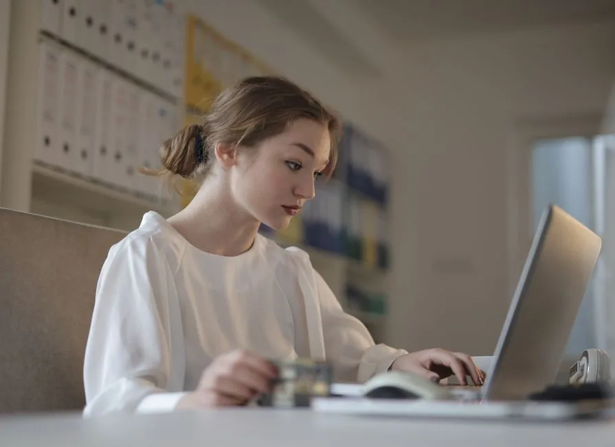 A small business owner working through her VAT return figures on a laptop, with accounts binders shelved behind her A small business owner working through her VAT return figures on a laptop, with accounts binders shelved behind her