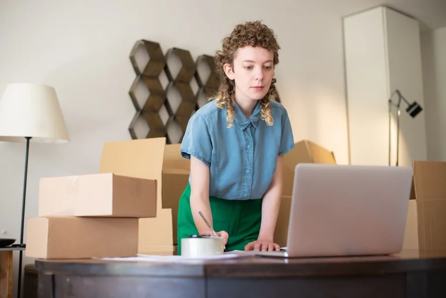 An online shop owner going through her paperwork at a laptop beside stacked stock parcels, working out her income tax bands. An online shop owner going through her paperwork at a laptop beside stacked stock parcels, working out her income tax bands.