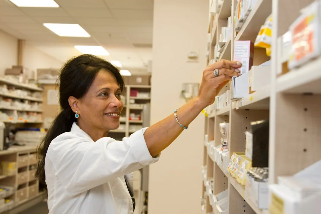 A locum pharmacist checking stock behind a community pharmacy counter while reviewing paperwork