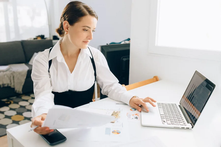 A UK small-business owner reviewing business finances on a laptop while holding a printed report at a desk A UK small-business owner reviewing business finances on a laptop while holding a printed report at a desk