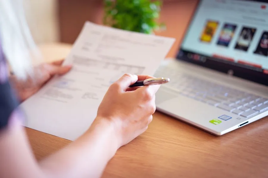 A landlord checking rental income paperwork against digital records on a laptop while managing tax alongside a job A landlord checking rental income paperwork against digital records on a laptop while managing tax alongside a job