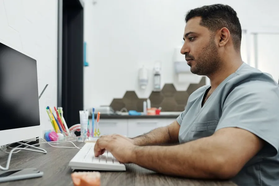 A dentist in scrubs working at a practice computer, reviewing the accounts behind the twice-yearly tax bill A dentist in scrubs working at a practice computer, reviewing the accounts behind the twice-yearly tax bill