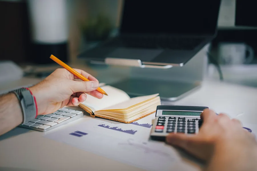 Payroll professional using a calculator beside a laptop and open notebook to check figures against a printed chart Payroll professional using a calculator beside a laptop and open notebook to check figures against a printed chart