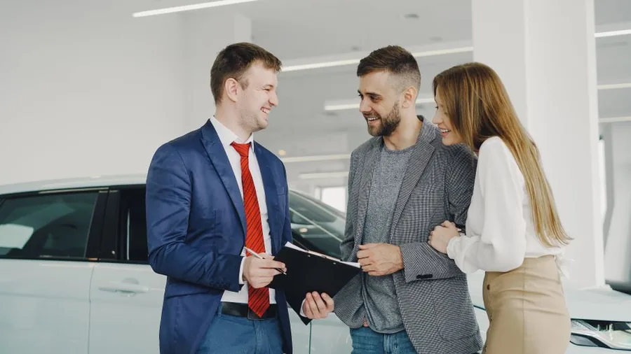 A car dealer talking a couple through the paperwork on a lease agreement beside a new car in the showroom A car dealer talking a couple through the paperwork on a lease agreement beside a new car in the showroom