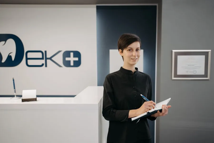A dental practice manager reviewing paperwork at a modern clinic reception beneath the firm's framed certification A dental practice manager reviewing paperwork at a modern clinic reception beneath the firm's framed certification