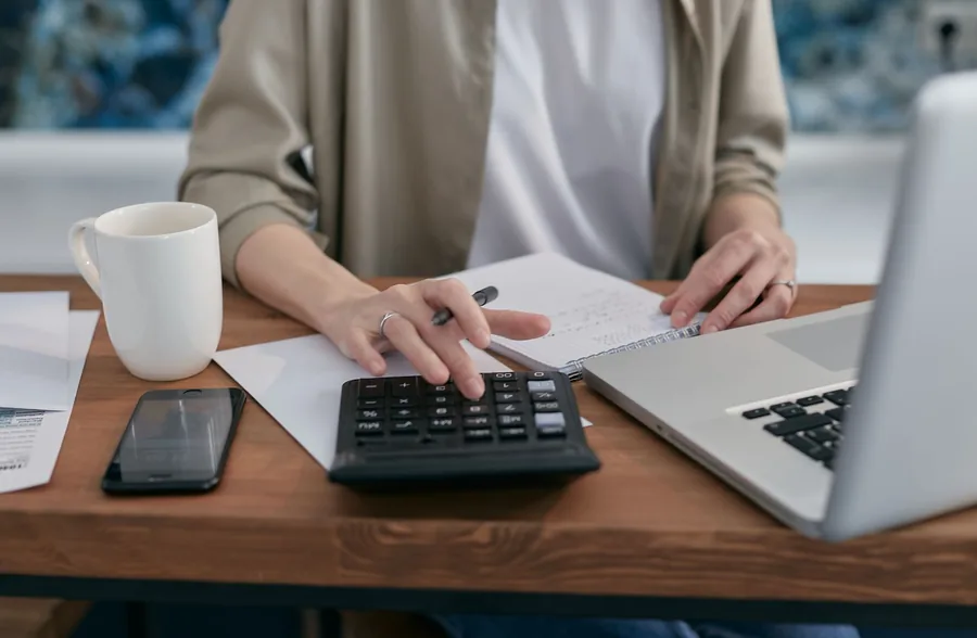 Self-employed worker at a wooden desk using a calculator and laptop while jotting figures to plan tax payments Self-employed worker at a wooden desk using a calculator and laptop while jotting figures to plan tax payments