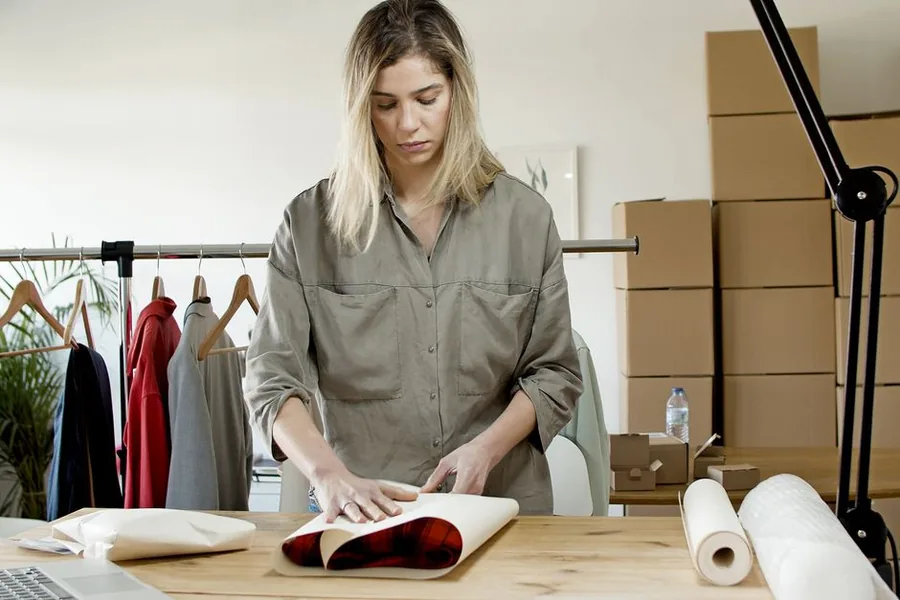 An online clothing seller wrapping stock to dispatch from a small ecommerce studio surrounded by shipping boxes