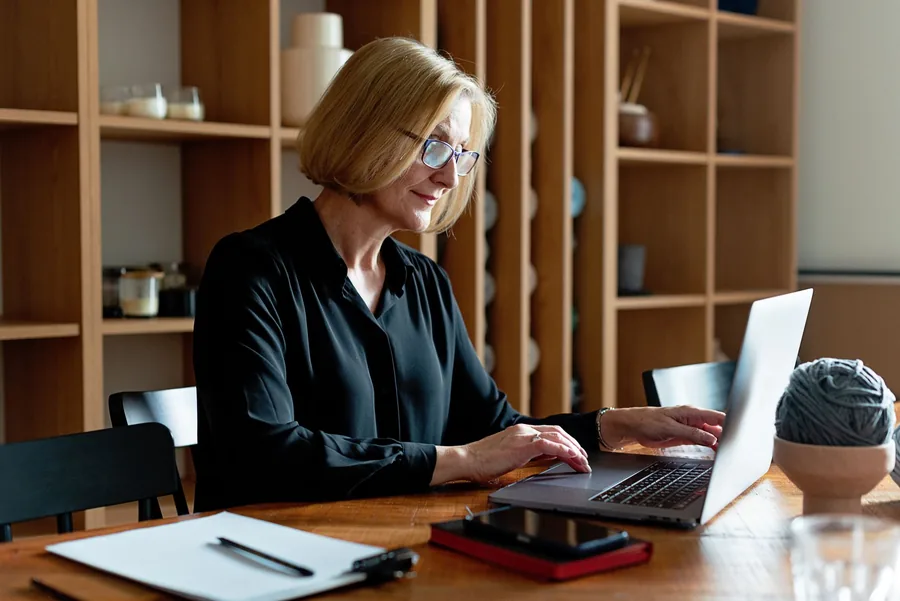 A woman in her fifties working calmly at a laptop at a tidy wooden table, a blank notebook beside her, checking her finances in warm daylight