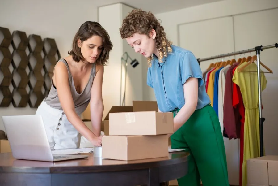 Two founders of a small online clothing business packing customer orders beside a laptop used to track their trading income