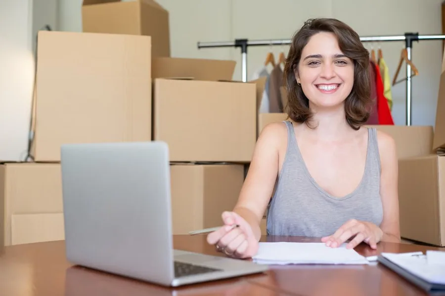 An online marketplace seller working at a laptop amid parcel boxes and a stock rail, reviewing sales records An online marketplace seller working at a laptop amid parcel boxes and a stock rail, reviewing sales records