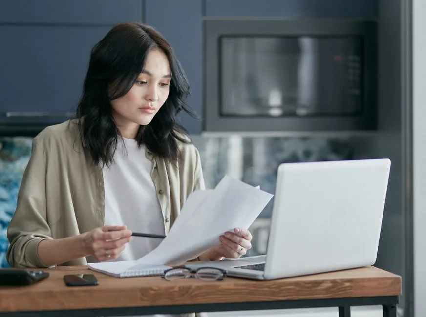 A business owner checking her VAT registration details on a laptop while holding a printed document at home A business owner checking her VAT registration details on a laptop while holding a printed document at home