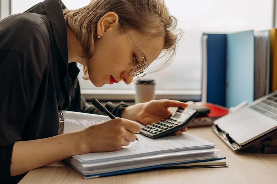 A bookkeeper reconciling ecommerce sales figures with a calculator and ring binders, separating gross takings from net A bookkeeper reconciling ecommerce sales figures with a calculator and ring binders, separating gross takings from net